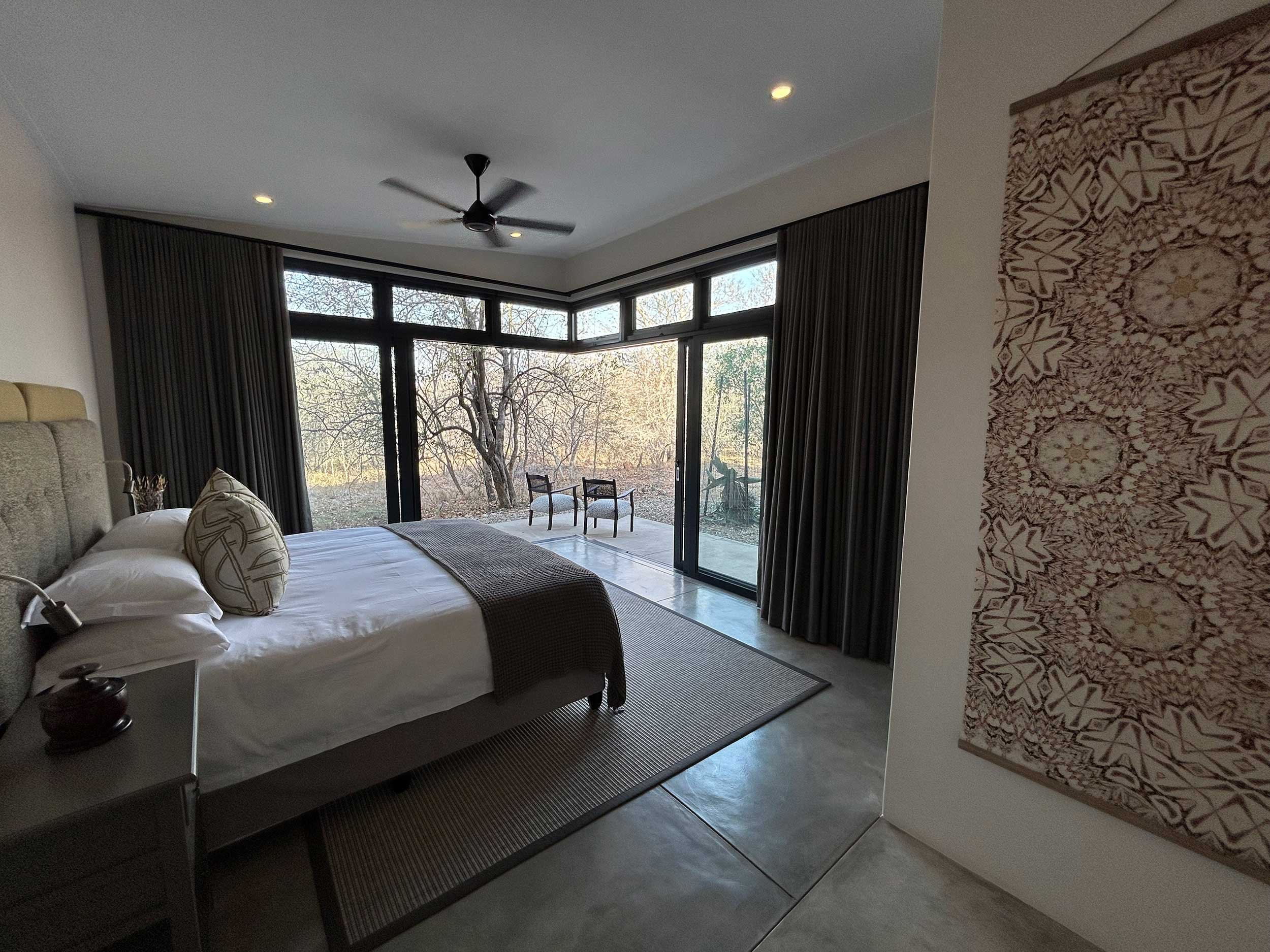 Modern bedroom with large windows, a bed with decorative pillows, dark curtains, and a patterned wall hanging. Outside the windows, there is a view of trees and a patio with chairs.