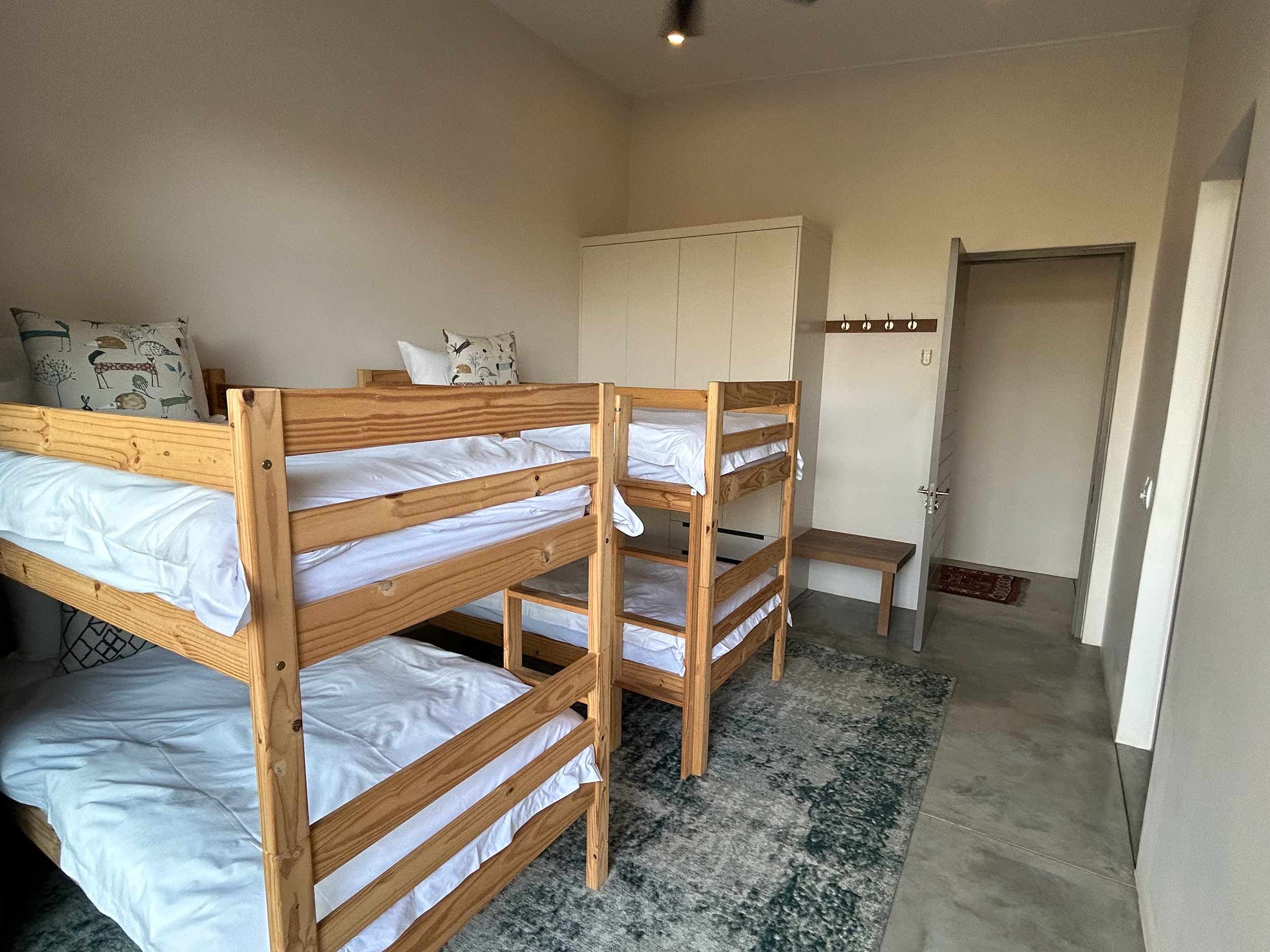 A clean, minimalist bedroom with two sets of wooden bunk beds, white bedding, decorative pillows, a wardrobe, a bench, and a small carpet on a concrete floor.