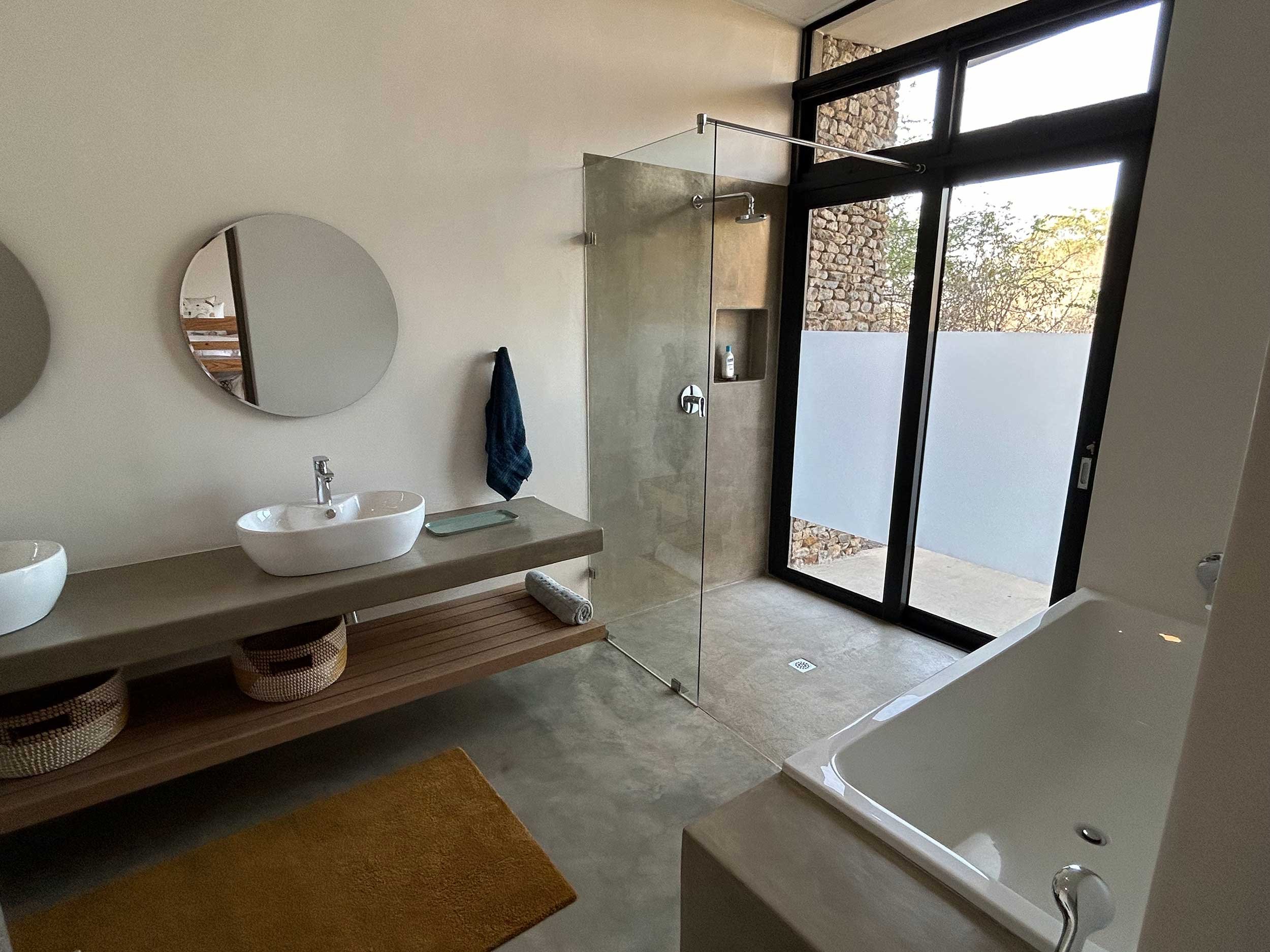 Modern bathroom with large mirror, dual sinks, glass shower enclosure, bathtub, and a tall window. Minimalist decor with beige and neutral tones.