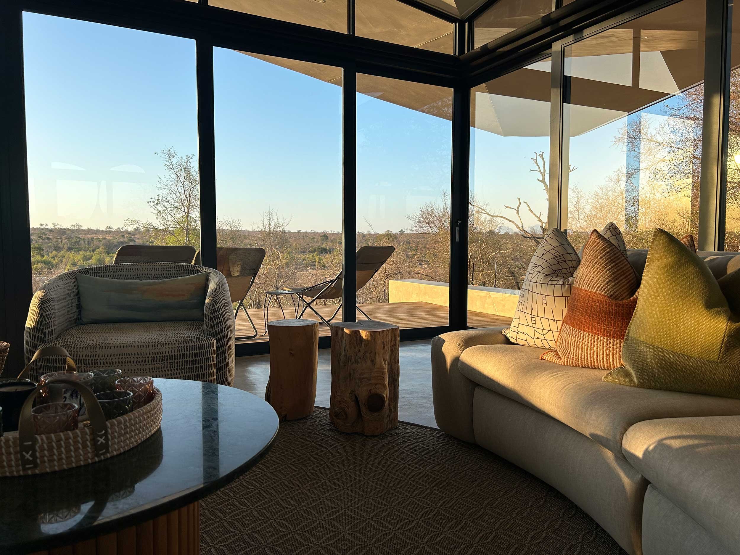 A cozy living room with modern furniture, including a wicker chair, round coffee table with a tray, and a sofa adorned with pillows. The room features large glass windows offering a view of a natural landscape with trees and clear skies.