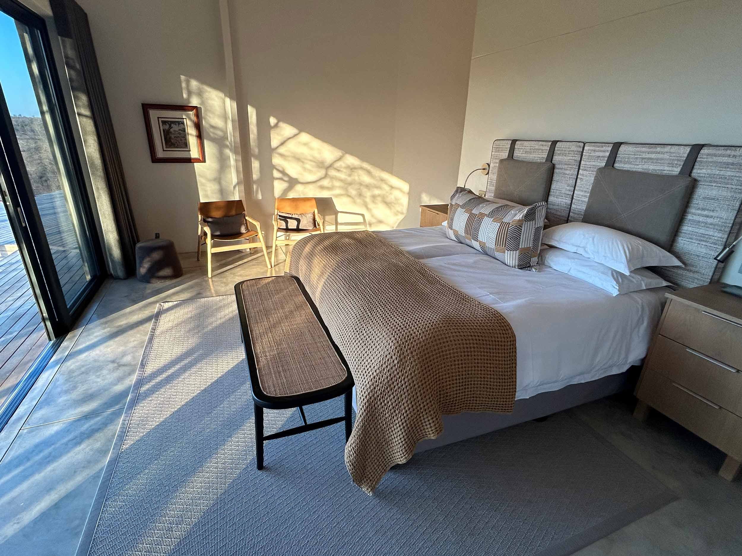 Modern bedroom with a large bed, beige bedding, and decorative pillows. A wooden bench sits at the foot of the bed. Two chairs are near a large window with a view of a deck. Soft light casts shadows on the walls and floor.