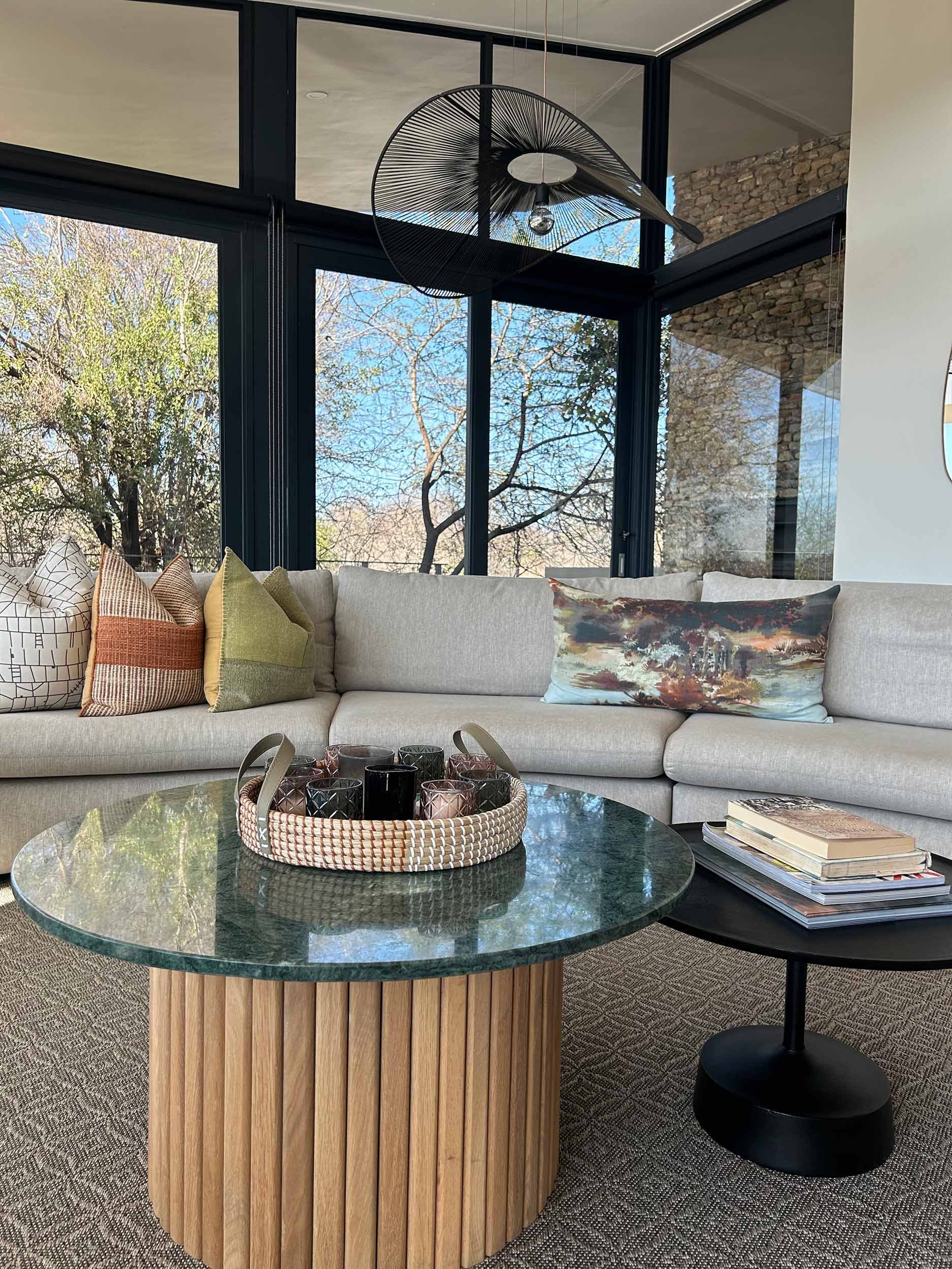 Modern living room with a light gray sectional sofa, colorful throw pillows, glass and wood coffee table with candles in a tray, a small black side table with books, large windows with a view of trees, and a contemporary ceiling light fixture.