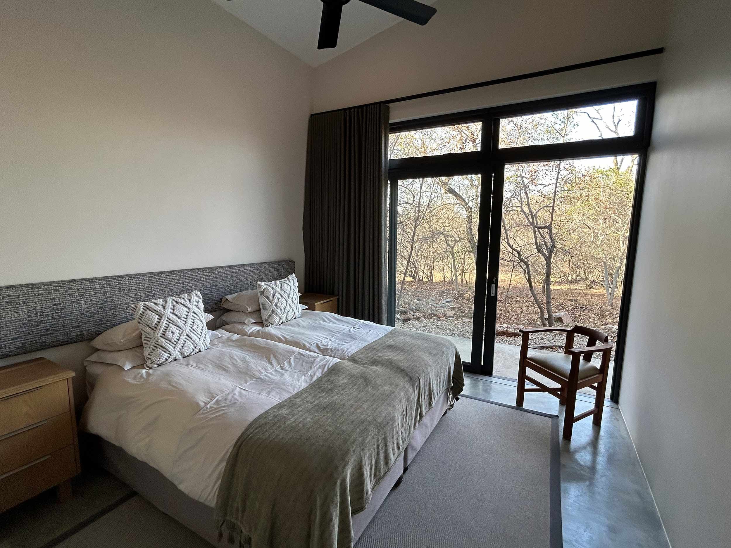 Minimalist bedroom with twin beds, wooden bedside tables, and a large window with a view of trees outside. A chair is placed next to the window, and the beds have white linens and patterned pillows.